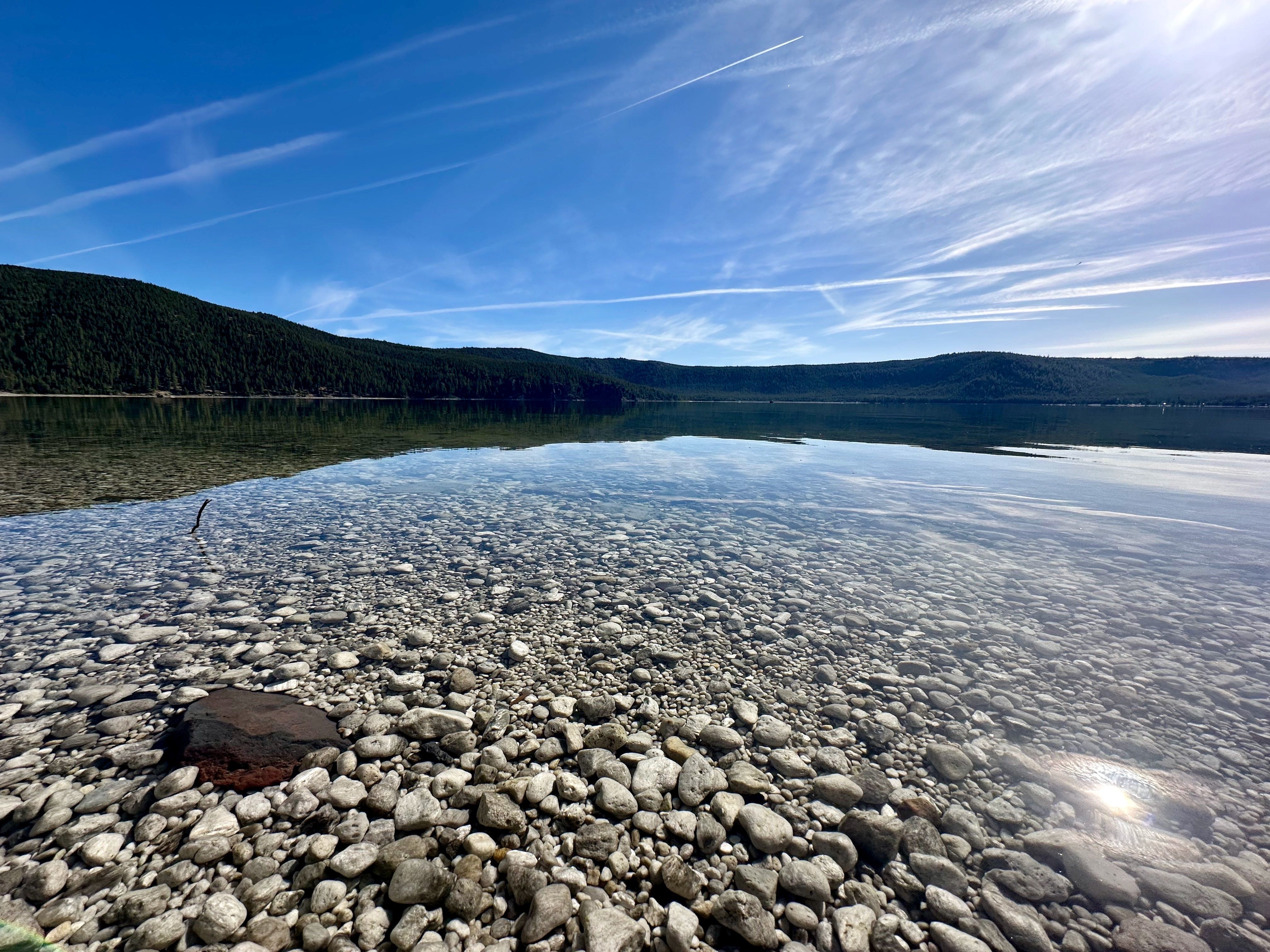 Lake with clear water, rocky shore, and mountains under blue sky.